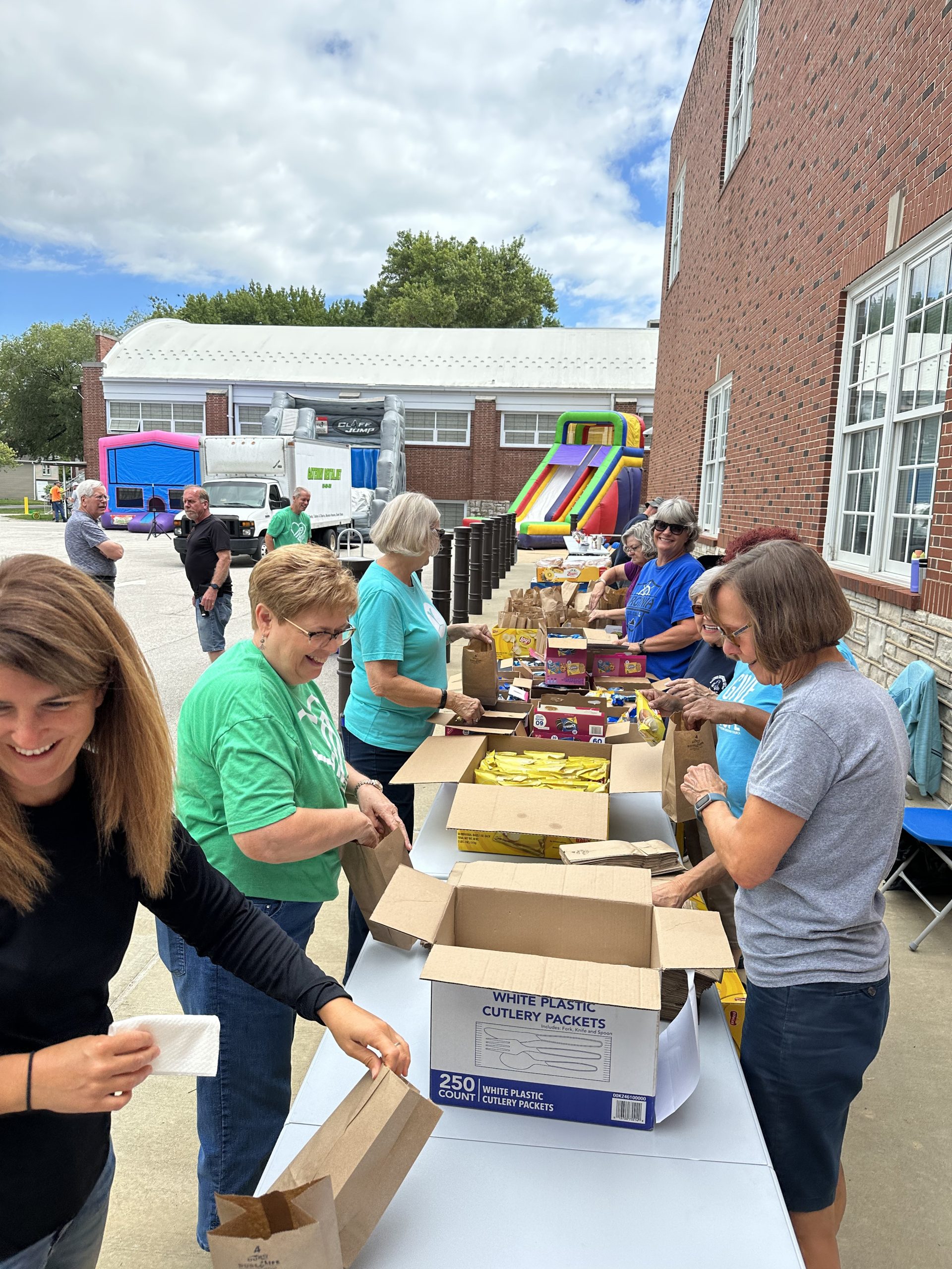 Volunteers in green and teal shirts packing lunch bags at an outdoor school Back to School Bash, with bounce houses behind