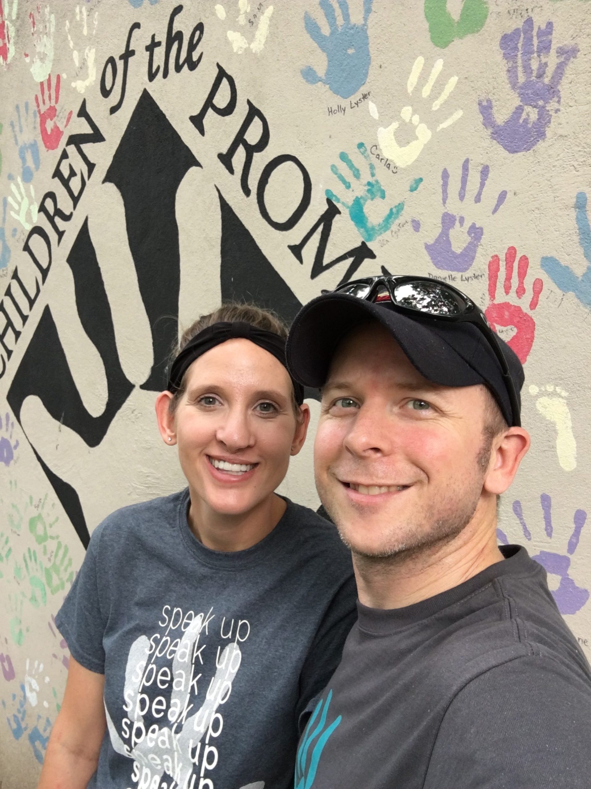 Mike Coumerilh and his wife smiling in front of a Children of the Promise wall covered in painted handprints