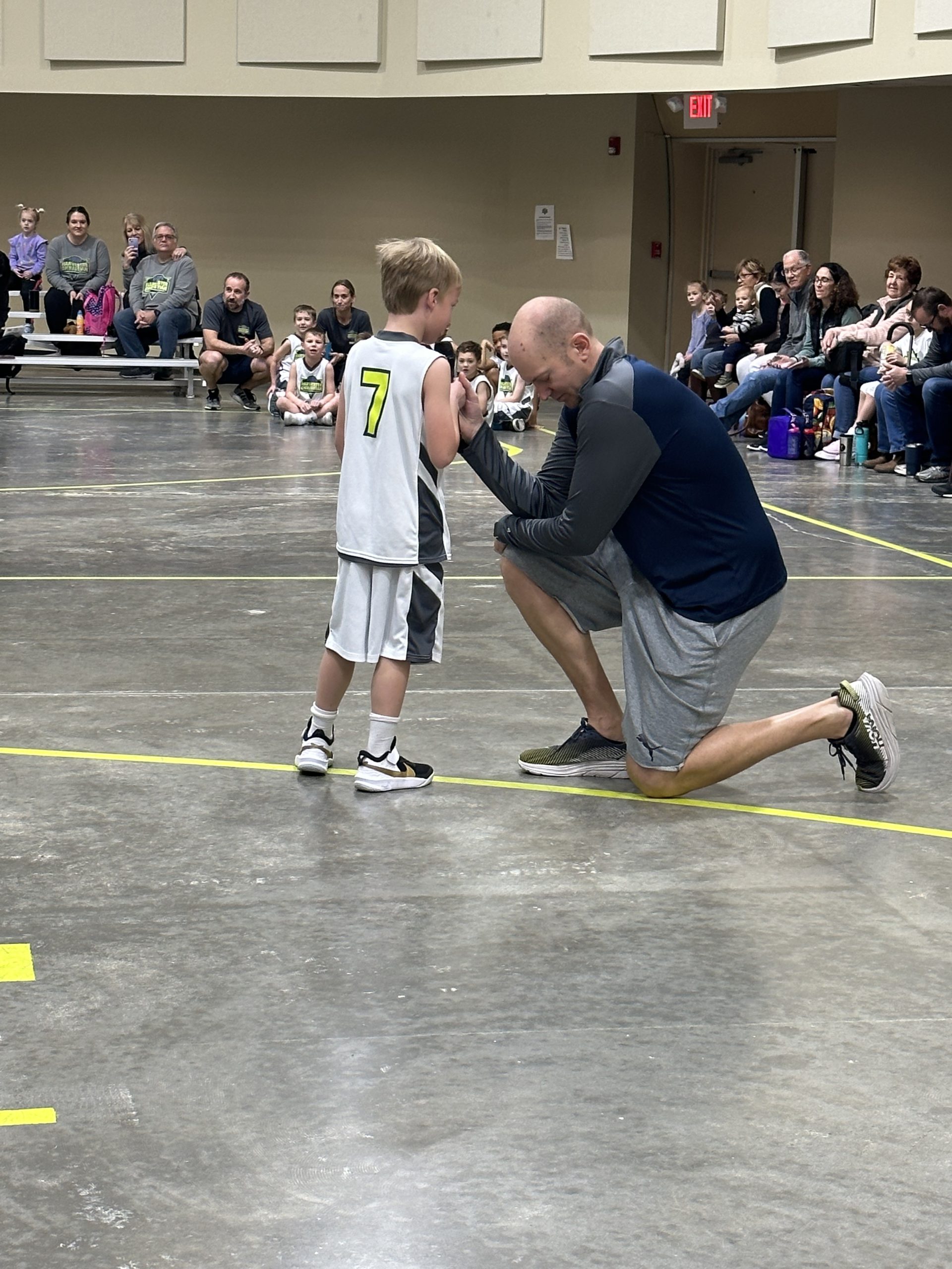 Coach kneeling to pray with a young basketball player in a number 7 jersey on the court as families watch