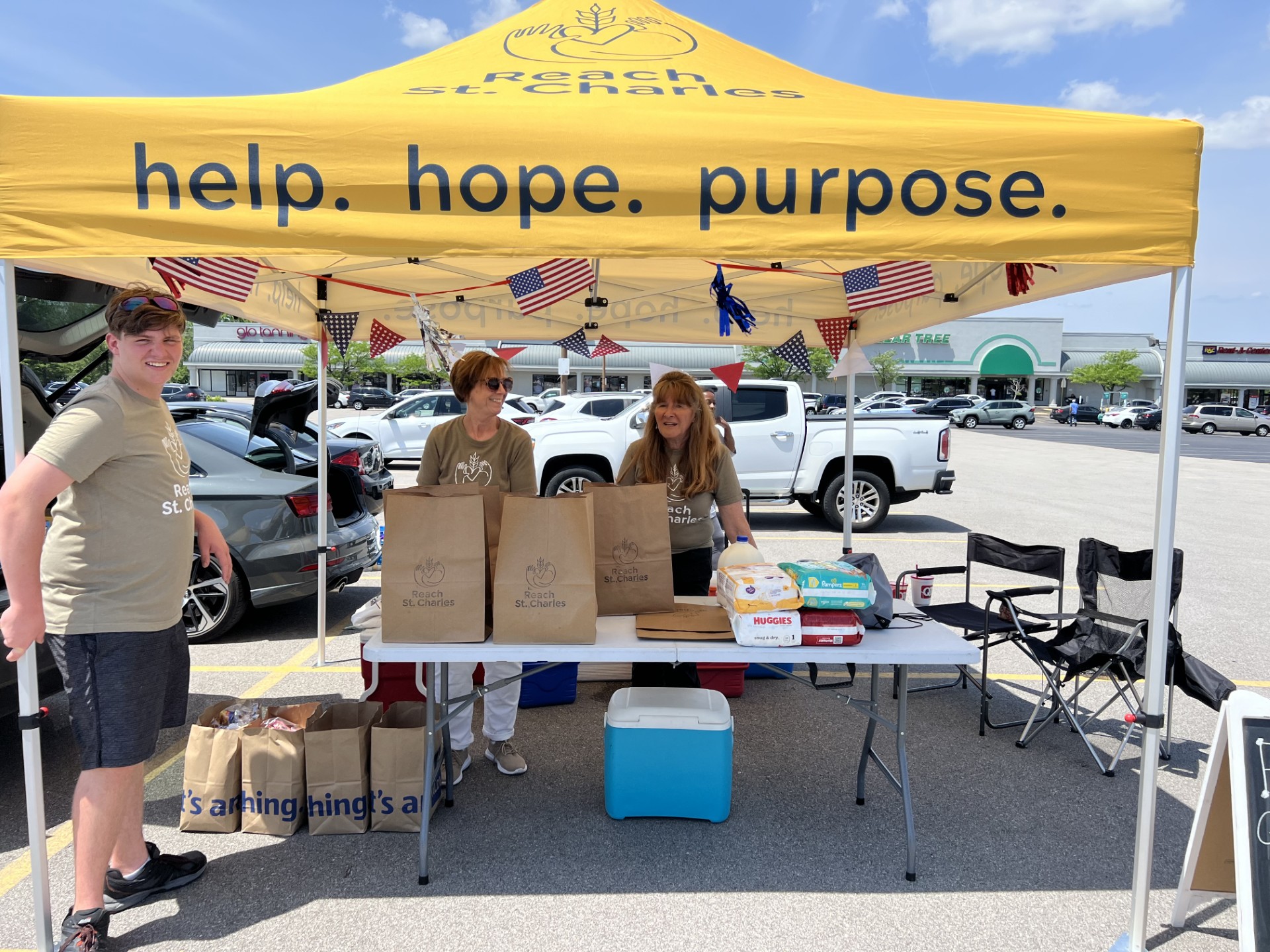 REACH St. Charles volunteers under a yellow 'help. hope. purpose.' canopy in a parking lot, with bagged food and diapers on a table