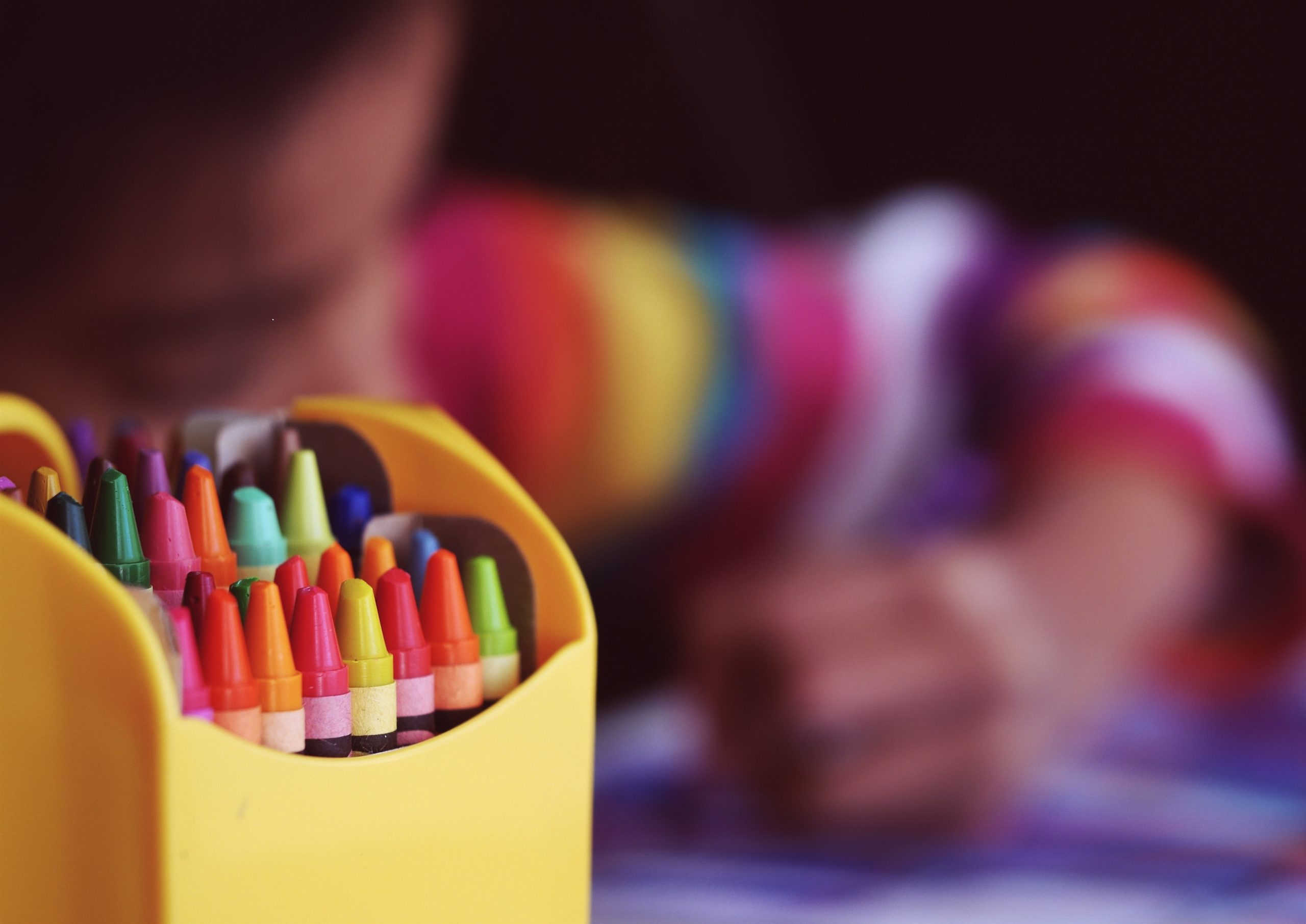 Box of colorful crayons in the foreground with a child drawing blurred in the background