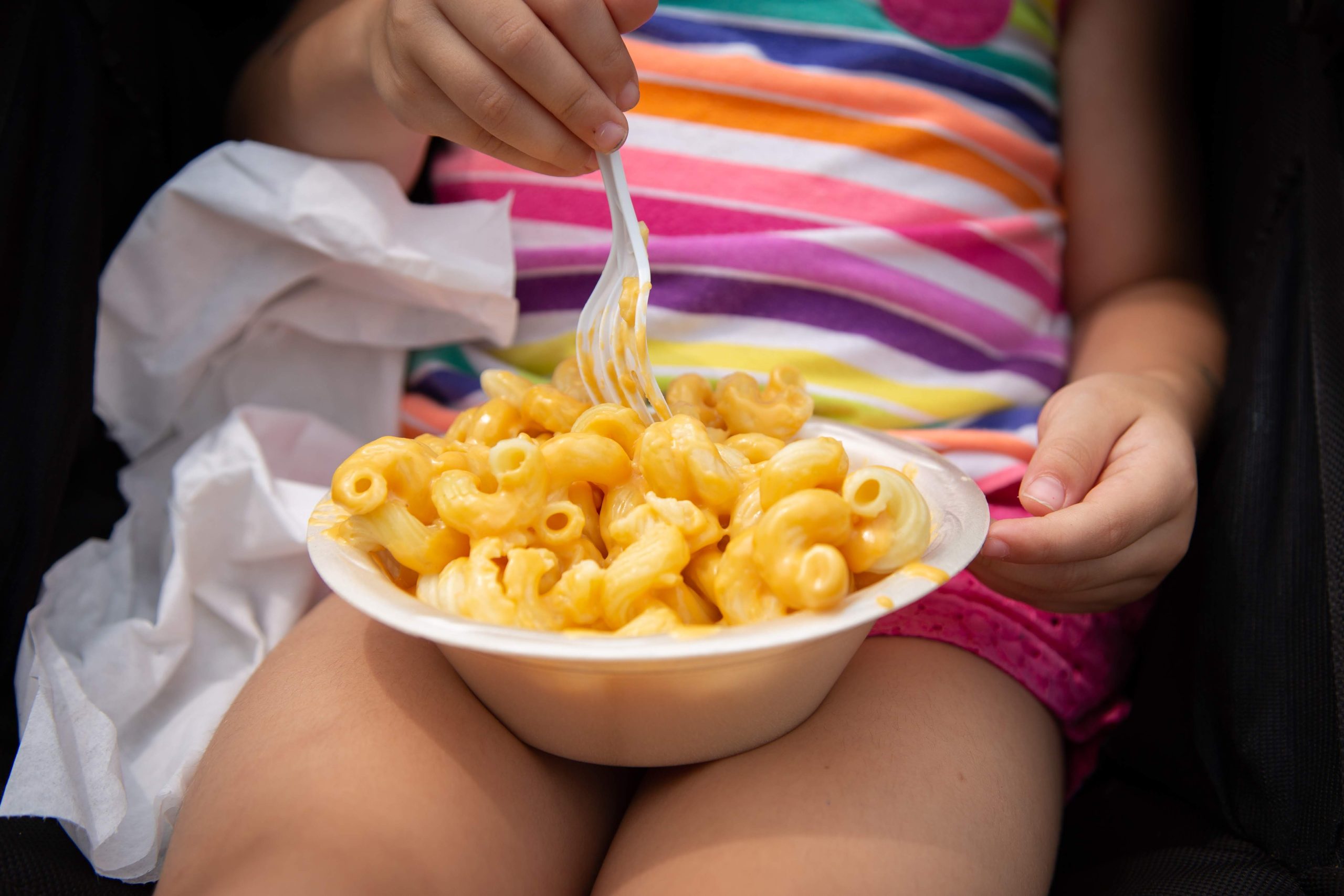 Child in a rainbow striped shirt eating macaroni and cheese from a paper bowl