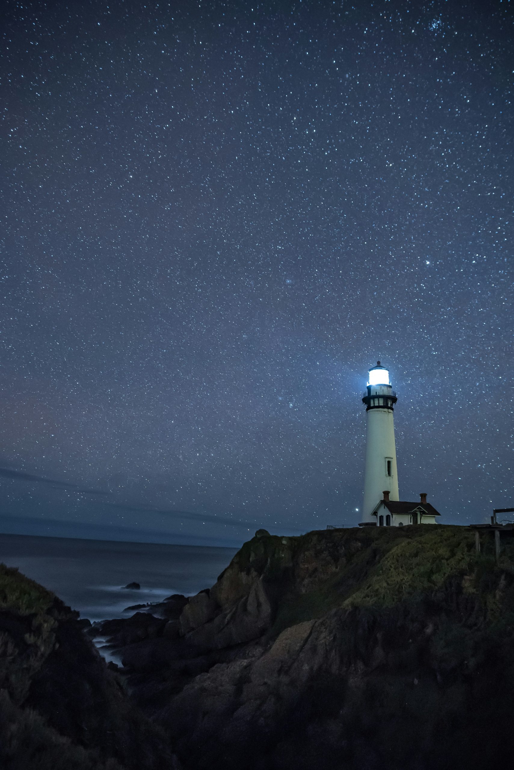 A lighthouse shining on a rocky coast under a star-filled night sky